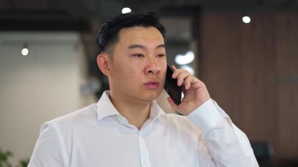 在公司的办公室里 一个有着严肃的面部表情的亚洲商人在用智能手机交谈 穿着白衬衫的成功人士与客户讨论约会的时间和日期 — 图库视频影像