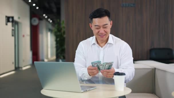 成功的男性雇主穿着白衬衫 坐在办公室桌上的现代笔记本电脑前 数着100美元钞票 快乐的亚洲人对公司的财务收入感到满意 — 图库视频影像