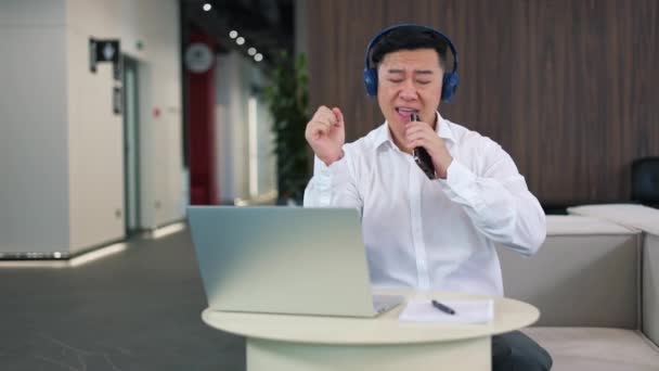 亚洲人坐在办公桌前 手持现代笔记本电脑 头戴无线耳机 用智能手机唱歌 就像麦克风一样 快乐的上班族们 享受着他们喜爱的音乐 在喝咖啡休息的时候享受着乐趣 — 图库视频影像