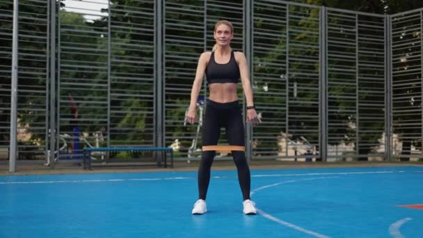 身穿深色运动服的精力充沛的女运动员蹲着 球场上有弹性的膨胀器 决心健身女士使用橡胶设备有效地训练和加强光溜溜和腿部肌肉 — 图库视频影像