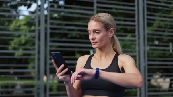 フィットネストラッカーアプリを使用してスポーツ服の若いアクティブな女性は 屋外で運動した後 彼女のトレーニングの進捗状況を監視します ブロンドの女性はスマートウォッチを見てから携帯電話で喜んで良い結果 — ストック動画