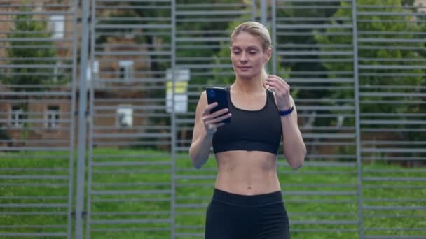 スポーツの若い女性は 屋外を歩き 耳にワイヤレスイヤホンを挿入します トレーニングを開始するための個人的なスマートフォンで音楽をオンにするワークアウト服を着たフィットとスレンダーの女性 — ストック動画