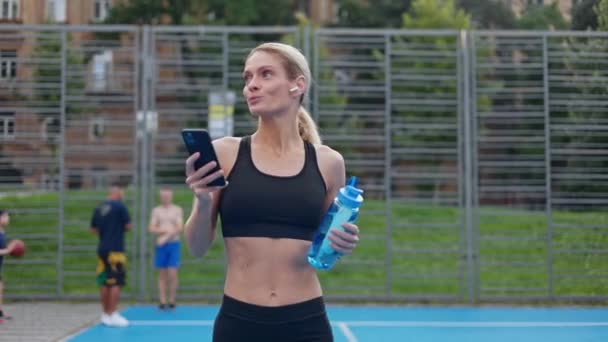 ブルーウォーターボトルで裁判所を歩き 音楽を楽しむときの満足感を表現する素晴らしい体型の運動女性 トレーニング中にスマートフォンやリモートイヤホンを使用して魅力的な女性 — ストック動画