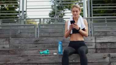 Açık renk saçlı sporcu kadın stadyum tribünlerinde dinleniyor ve akıllı telefondan mesaj atarken gülümsüyor. Spor kıyafeti giyen genç bir sporcu sağlıklı bir yaşam tarzı ve açık havada antrenman yapıyor..