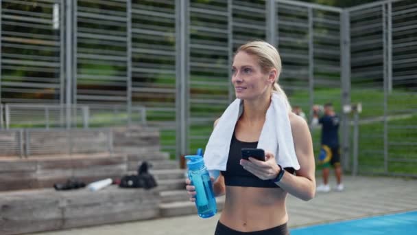 身穿黑色运动服 身材苗条的健身女士在球场上行走 从成功的户外运动中获得满足感 用蓝色水瓶微笑的女人在手机上看了一眼 — 图库视频影像