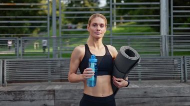 İnce beyaz kadın elinde içecek şişesi ve eskrimli spor sahasının arka planında bükülmüş siyah yoga minderiyle poz veriyor. Kaslı vücutlu gülümseyen kadın dışarıda yoğun bir idmana hazırlanıyor..