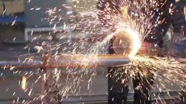 Koruyucu giysili ve eldivenli, uzun boruları öğütücüyle kesen çalışkan bir erkek. Alevli kıvılcımlar farklı yönlere uçarken metali iki parçaya bölen özel bir araç.