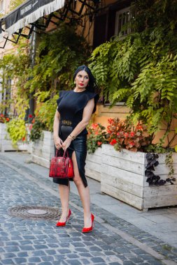 Siyah bluz, etek ve kırmızı topuklu ayakkabı giyen kendine güvenen bir iş kadınının uzun bir portresi kaldırım taşı şehir caddesinde elinde moda çantasıyla dikiliyor. Beyaz kadın düşünceli bir şekilde yan tarafa bakıyor..