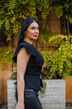 Zarif siyah bluz ve deri etek giyen başarılı iş kadınının yan görüntüsü yeşil tırmanma tesisleri olan eski binaların yakınında geziniyor. İnsanlar ve yaşam tarzları kavramı.