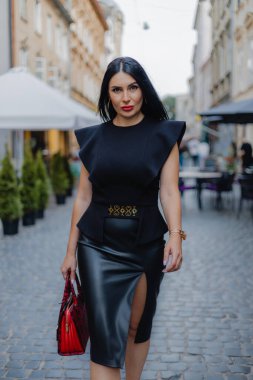Uzun siyah saçlı, kırmızı dolgun dudaklı, çekici orta yaşlı bir kadın model yürüyüşü ile caddede yürüyor. Siyah bluz ve deri etek giyen kendine güvenen iş kadınının açık hava portresi..