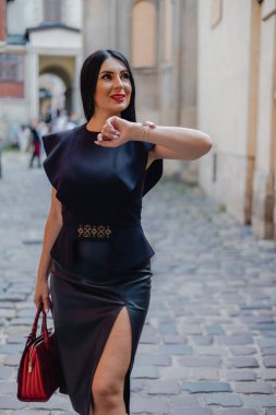 Eski şehrin kaldırım taşı sokağında duran deri etek ve siyah bluz giyen orta yaşlı zarif bir kadın. Çekici esmer, kırmızı çantalı, elini dirseğine sokmuş ve yana bakıyor..