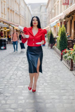 Orta alanda duran beyaz tenli çekici bir kadının elinde tabak ve bir fincan kahveyle görüntüsü. Zarif stil sahibi kadın antik şehrin sokaklarında yürüyor ve eğleniyor..