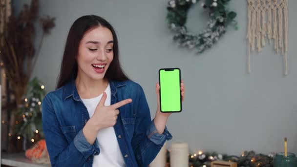 お祝いの装飾されたクリスマスキッチンの若い女性は 緑色のスクリーンでスマートフォンを提示し 親指を与えることを指摘し 新年の祝祭の広告 ビデオや写真 — ストック動画