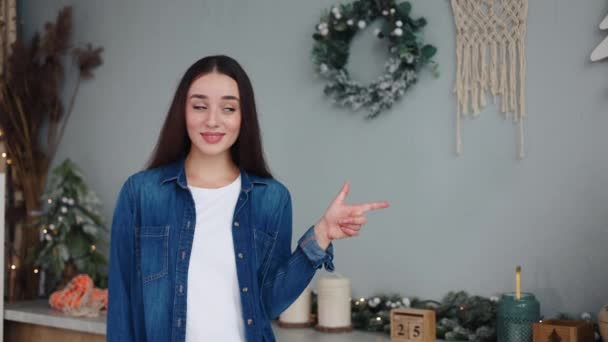 彼女のお祝いの装飾された家のキッチン ガーランドで飾られたクリスマスツリーのワークスペースモックアップエリアに向かって白いTシャツのジェスチャーの少女 プロモーションコンテンツやお祝いの広告のための完璧な背景 — ストック動画