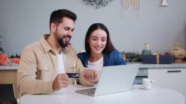 Mutlu çift Noel hediyeleri için mutfaktan online alışveriş yapıyor, mağazalara bakmak için dizüstü bilgisayar kullanıyor, yeni yıl hediyeleri için kredi kartıyla ödeme yapıyor. Mükemmel kış tatili alışveriş günü..
