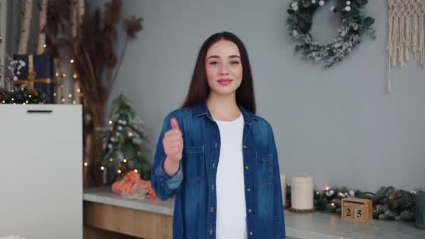 白いティーとブルーのシャツの魅力的な陽気な少女は サムスンアップジェスチャーを与えるお祝いの装飾されたクリスマスキッチンに立っています 女性はポジティブさを示し 承認を伝える 広告のための理想的な場所 — ストック動画