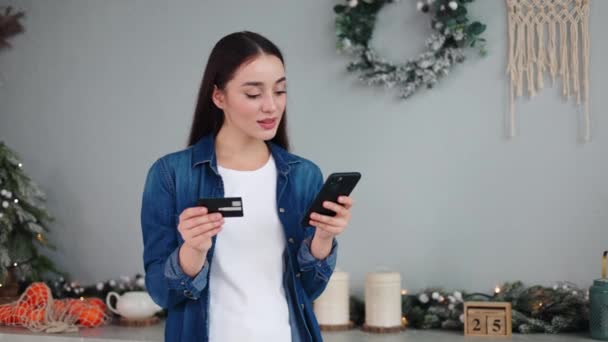 スマートフォンでクレジットカードコードを入力してクリスマスツリーの近くの自宅キッチンでワクワクする勝利を祝う笑顔の幸せな女性 新年の販売時間 Eコマースのウェブサイト — ストック動画
