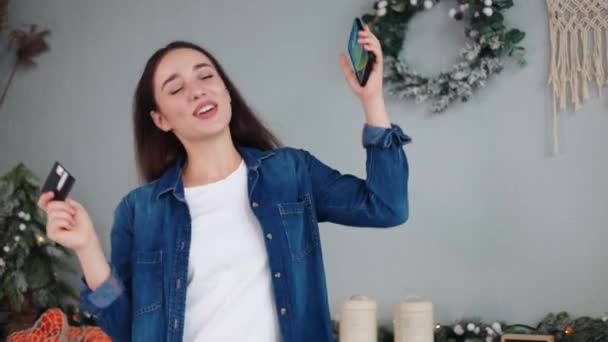 スマートフォンでクレジットカードコードを入力して新年のギフトを購入し クリスマスツリーの近くの自宅のキッチンで手を挙げてダンスを興奮させるウィンを祝う楽しい幸せな女性 新年の販売 Eコマースのウェブサイト — ストック動画