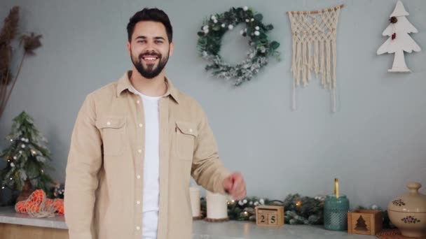 一个容光焕发的男人 带着灿烂的笑容和蓝眼睛的姿态 在一个装饰华丽的厨房里模仿空间 在节日广告和促销寒假的完美背景下照亮着圣诞树 — 图库视频影像