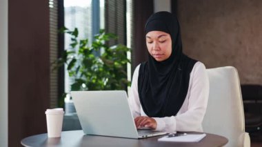 Çok kültürlü, siyah tesettürlü ve resmi kıyafetler giyen bir kadın ofis masasında oturuyor ve kablosuz bilgisayarda yazı yazıyor. Becerikli bayan yönetici odaklanmış yüz ifadesiyle ekrana bakıyor.