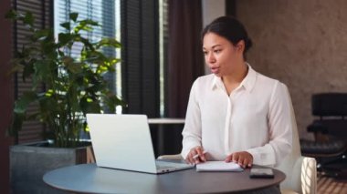Ofis masasında oturan ve video görüşmesi için kablosuz bilgisayarı kullanan odaklanmış bir kadın işçi. Beyaz bluzlu Afro-Amerikalı kadın elinde kalemle çevrimiçi toplantı sırasında bilgi yazıyor..