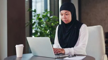 Resmi olarak giyinmiş bir bayan yuvarlak masaüstündeki taşınabilir bilgisayarda yazarken düşünceli bir şekilde yana bakıyor. İş yerinde iş mektubu yazarken doğru kelimeleri arayan tesettürlü çok kültürlü bir kadın..