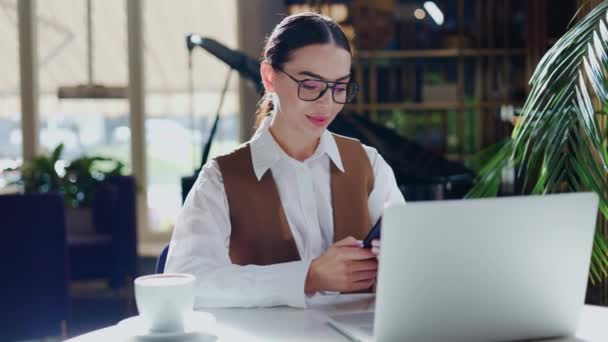戴眼镜 身穿时髦服装 坐在餐桌旁 在笔记本电脑上工作 一边用手机上网 一边抬头望着相机 一边笑着的富有梦想的 快乐的商界女性 — 图库视频影像