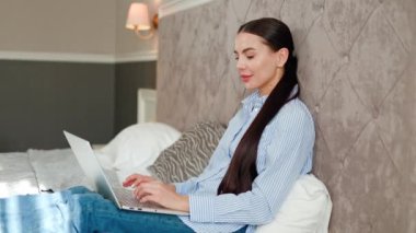 Otelde dizüstü bilgisayarda çalışan, yatakta oturan, iş gezisi sırasında ışıl ışıl gülümseyen güzel başarılı iş kadını. Mesafe çalışması, iş.