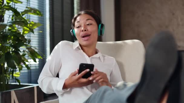 ハッピービジネスレディーは 仕事の日に休んでいる間 快適なオフィスの椅子で歌ったり踊ったりします オフィスで楽しむためのワイヤレスヘッドフォンとモダンなスマートフォンを使用してアフリカ系アメリカ人女性 — ストック動画