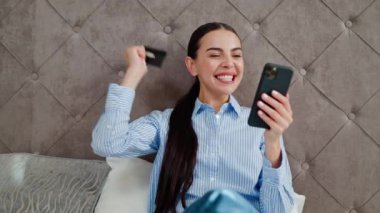 Cep telefonuyla video oyunu oynayan heyecanlı bir kadın, iyi haberler okuyor, kredi kartı tutuyor, piyangoyu kazanmayı kutluyor, mutlu ve neşeli bir jest yapıyor, otel odasında oturuyor..