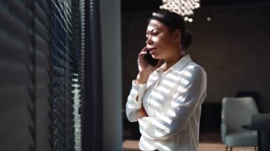 Şirketin iş yerinde duran ve modern akıllı telefonu sohbet için kullanan çok ırklı kadın yöneticinin yan görüntüsü. Telefon görüşmesi sırasında pencereden bakan afro saçlı güzel bayan..