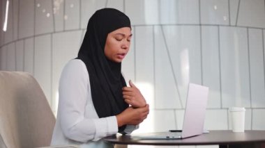 Kablosuz bilgisayarda sürekli çalışmanın yol açtığı bilek ağrısı çeken tesettürlü Afrikalı Amerikalı bir kadın. Modern işyerinde oturan yorgun kadın ve masaj yapan eller. Ofis sendromu ve sağlık hizmetleri kavramı.