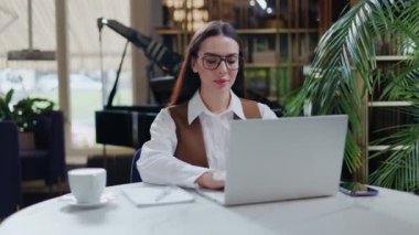 Yoğunlaşmış iş kadını modern dizüstü bilgisayarın önünde yuvarlak masada oturuyor, ekrana bakıyor ve internetten bilgileri not defterine kopyalıyor. Teknoloji ve mesafe çalışma görevi kavramı.