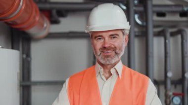 Gri saçlı fabrika işçisinin kollarını kavuşturup kameraya gülümsemesini yakından izleyin. Yelekli ve kasklı beyaz adam endüstriyel aktivitelerde başarıyı temsil ediyor..