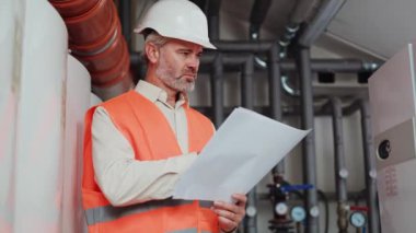 Koruyucu kask ve yelek giyen, atölyenin üzerinde duran ve planlara bakan, ehil bir inşaat işçisi. Beyaz sakallı erkek, kapalı alanda kağıt üzerinde bilgi kontrol ediyor..