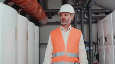 Portakal rengi güvenlik yeleği giyen gururlu erkek mühendis atölyenin arka planında dizüstü bilgisayarı kapalı duruyor. Yakışıklı yaşlı erkek modern fabrikada kalite kontrolü sağladıktan sonra kameraya bakıyor..