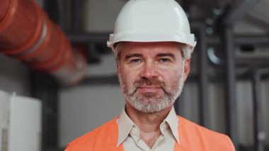 İş yerinde poz veren beyaz, turuncu yelek giyen bir fabrika çalışanının portresi. Gri sakallı ve saçlı inşaat mühendisi teknik odasındaki karmaşık işinden sonra göz teması kuruyor..