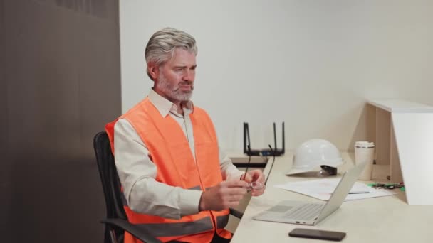 現代のラップトップの過労のために胸の痛みに苦しんでいる安全オレンジ色のベストを身に着けている灰色のハイレッド産業労働者 デスクに座り 職場で心臓発作を起こしている高齢の白人男性 — ストック動画