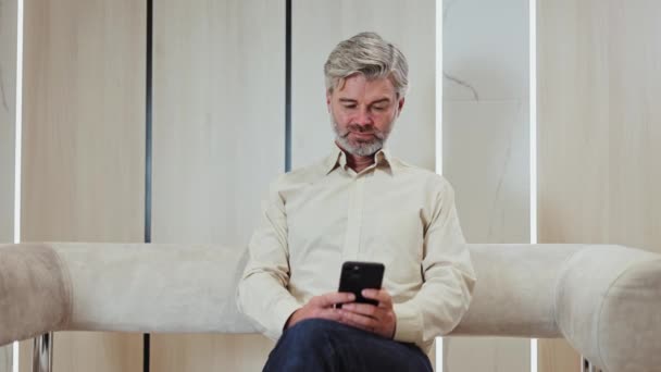グレイはオフィスで快適なラウンジチェアに座っている間 現代のスマートフォンにテキストを送信する男性をひげました 個人的なガジェットを使用して仕事の電子メールに答える正式な服を着ている白人高齢者 — ストック動画