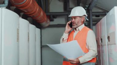 Sinirli fabrika işçisi modern akıllı telefondan konuşuyor ve elinde inşaat çizimleri tutuyor. Sakallı, kır saçlı erkek kavga ediyor ve teknik odada geliştirilmiş plandaki hataları tartışıyor..