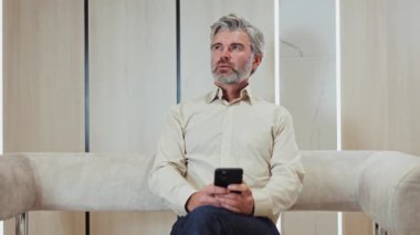 Resmi giyinmiş Kafkasyalı kıdemli işadamı, elinde modern akıllı telefonuyla rahat koltukta otururken düşünceli bir yüz ifadesiyle yan yana bakıyor. İş, teknoloji ve yaşam tarzları kavramı.
