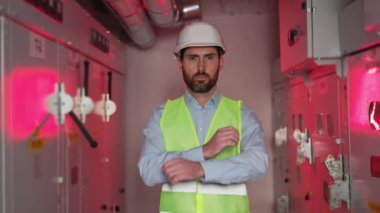 Santralde kollarını kavuşturan bir erkek teknisyen yeşil yelek, kask takmış, kırmızı arka plan, elektrik santrali ayarında tehlike arz ediyor. Profesyonel elektrikçinin portresi.