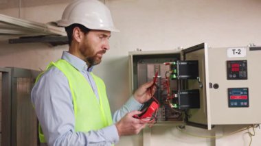 Elektrik testinde yetenekli bir elektrikçi elektrik santralinde santral işlevselliğini denetliyor, koruyucu kıyafet teknisyeni fabrika teknik alanındaki aktarım hatlarında voltajı inceliyor..
