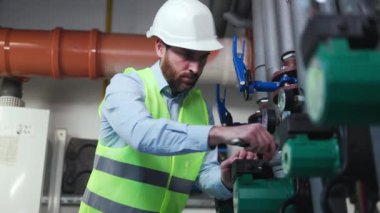 Koruyucu miğferli tesisatçı kazan dairesinde fındık sıkıyor. Tamirci, ısıtma sisteminin teknik alanında sıcak su borusunu tamir ediyor. Borulardan su akışını ayarlamak. Tesisat onarım servisi.
