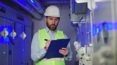 Koruyucu giysiler içindeki elektrik mühendisi elektrik santralindeki panoya not alıyor. Fabrikanın teknik odasındaki verileri ve ölçümleri kontrol eden adam. Dağıtım elektrik santralleri.
