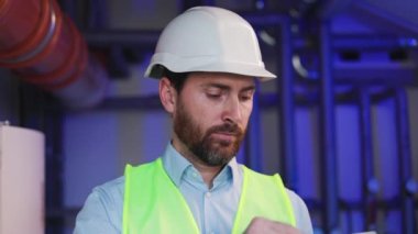 Atölye ortamında profesyonel tesisatçı elinde kablosuz bir dizüstü bilgisayarla mavi arka planda veri kaydediyor. Adam beyaz bir şapka takıyor ve kameraya bakıyor..