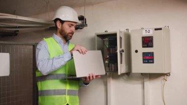 Elektrik mühendisi kontrol odasındaki elektrik dağıtım kabinesinde izleme işlemleri için dizüstü bilgisayar kullanıyor. Önleyici bakım yılı, şirkette çalışan profesyonel elektrikçi..