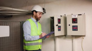 Erkek işçi, bir metalurji fabrikasının makine bölümündeki verileri analiz ediyor. Elektrikçi, elektrik odasındaki bir dağıtım panosundan gelen verileri bir tablete kaydediyor. Elektrik ekipmanları.