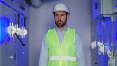 Elektrik santralinde beyaz şapka ve üniforma giyen yetişkin bir elektrikçinin portresi. Üniformalı ve kasklı başarılı bir erkek inşaat yöneticisi mavi arka plandaki kameraya bakıp gülümsüyor..