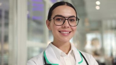 Hastane çalışanlarının mutlu portresi gözlük takıyor ve hastanede gülümserken ayakta duruyor. Kafkasyalı mutlu kadın doktor kapalı alanda başarılar ve büyük başarılar gösteriyor..